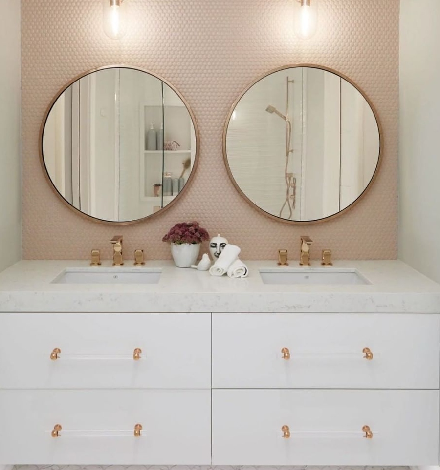 Bathroom designs ideas by Dvira Interiors. The bathroom featured in this image is a great representation of symmetry in design. The use of rose gold brass fixtures is particularly eye-catching with the warm blush tiled backsplash. The round mirrors offset the rectangular sinks and cabinetry below. Looking closely, the scalloped floor tiling adds a further dimension to this bathroom. ⁠