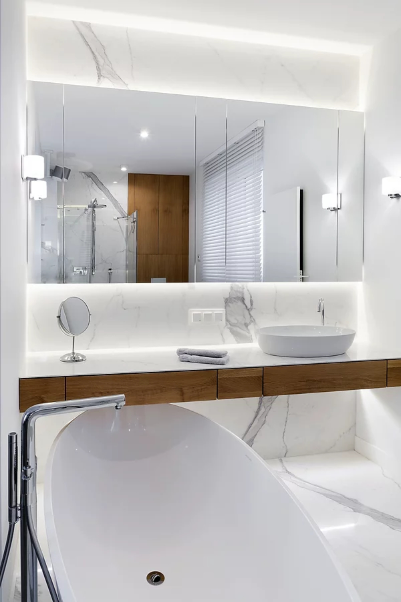 apartment p bathroom with an oval bathtub, a wood wash basin with a white sink. A long rectandular mirror and some wall lamps. It also has a marble surface.