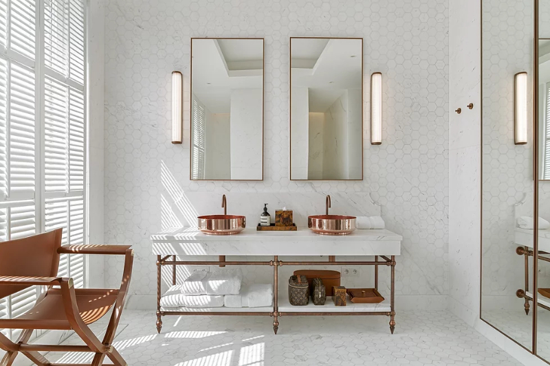 apartment k master bathroom with 2 sinks in rose gold, a brown chair, and 2 lamps next to the square mirros.